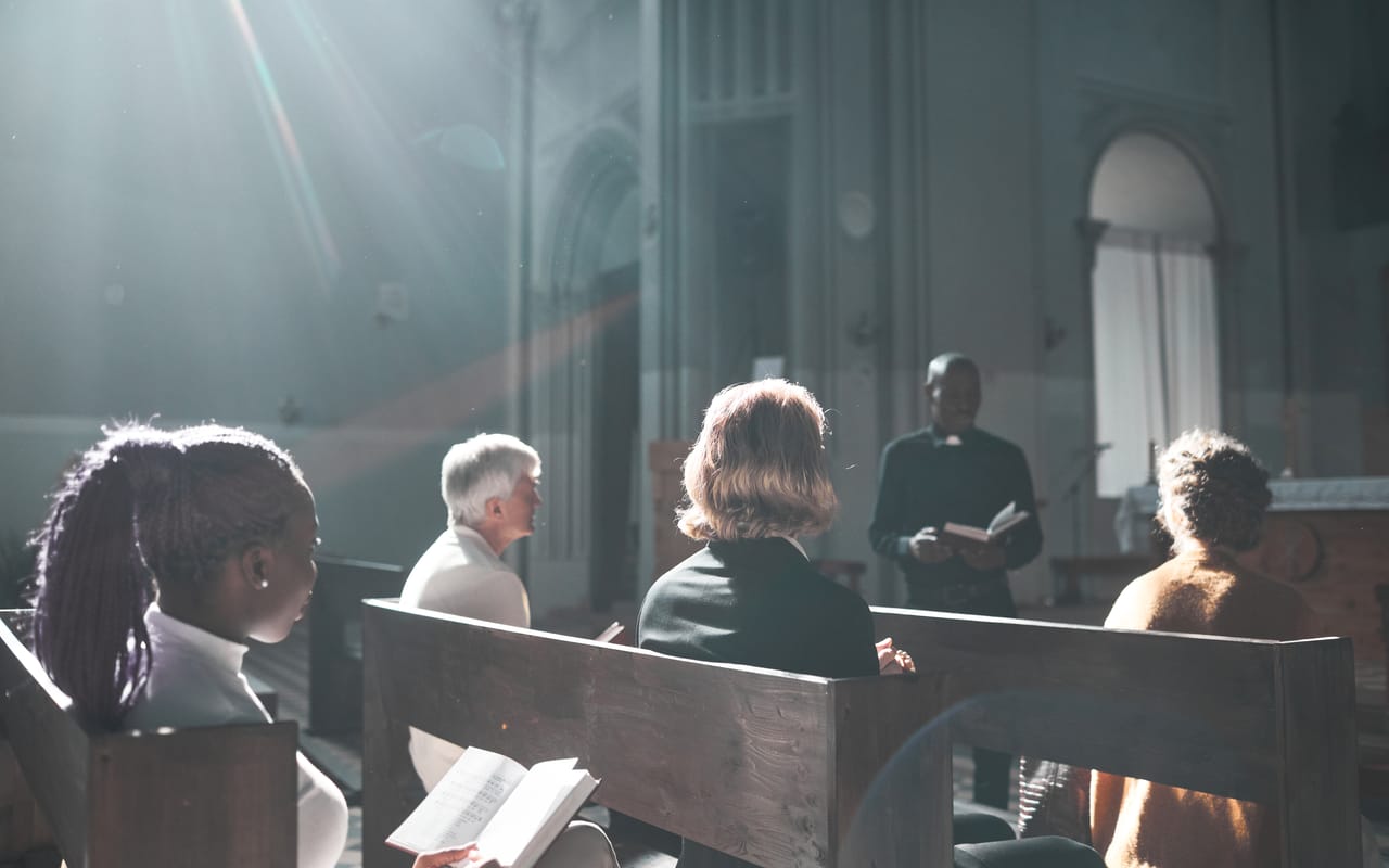 People seated in church pews as light falls across them, a pastor reading at the front
