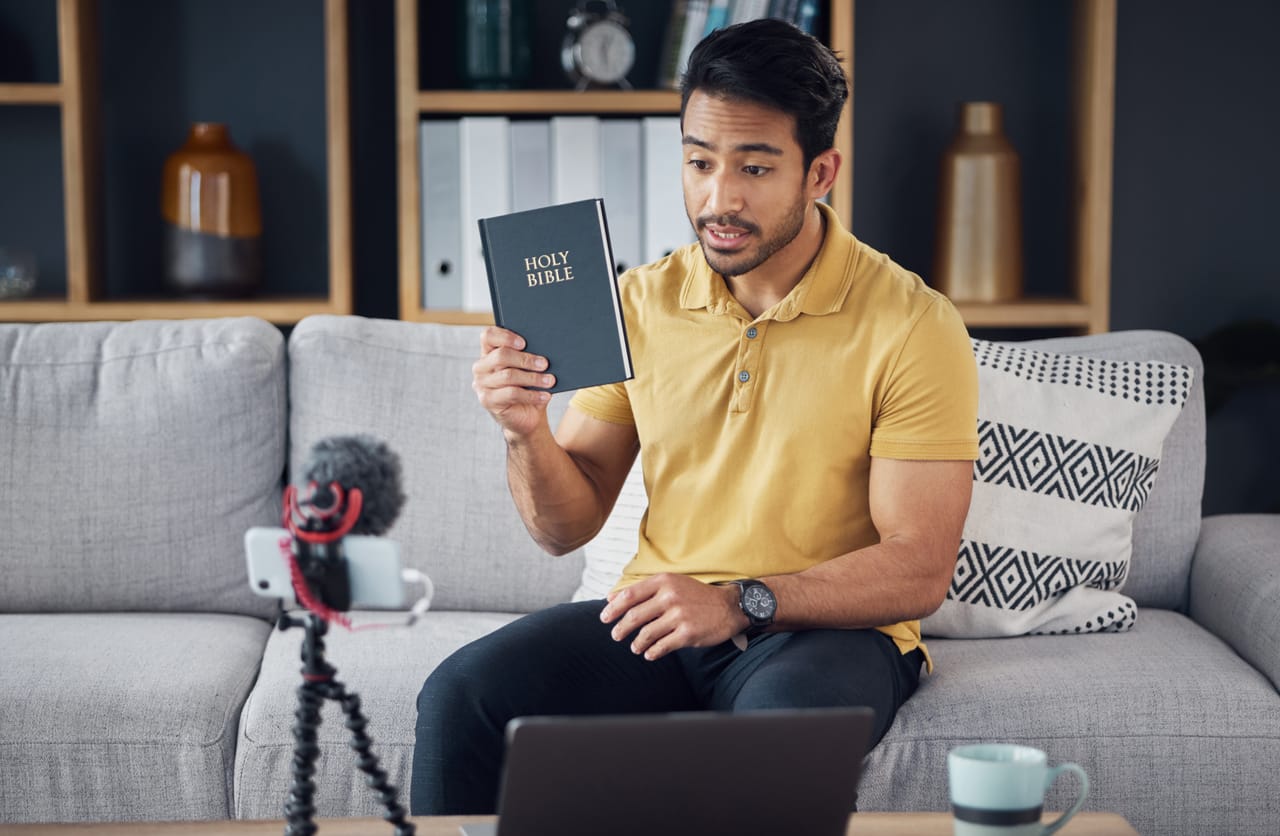 A man on a sofa holding up a Bible to a phone camera during a video call