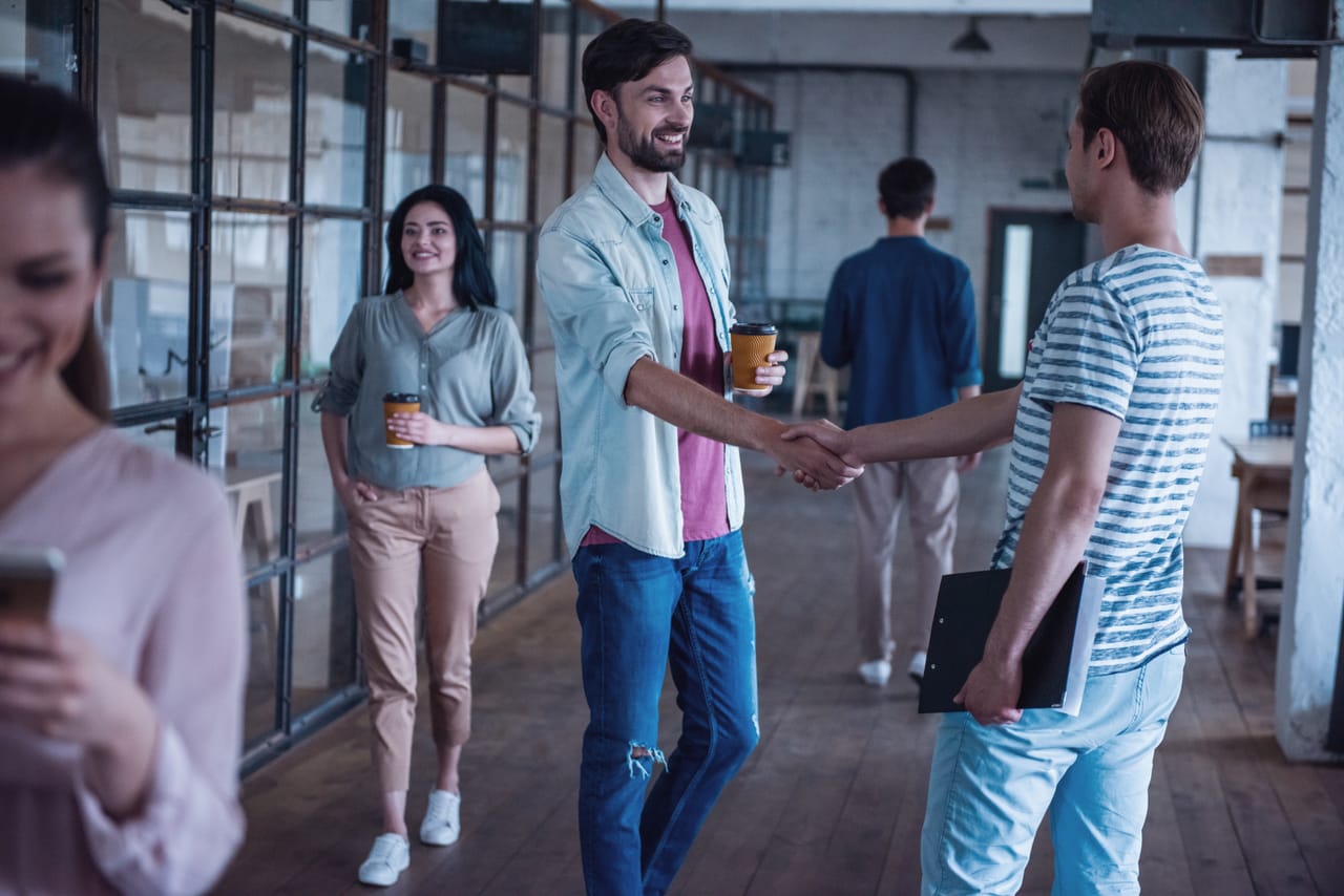 Two people shaking hands as others arrive with coffee before a gathering
