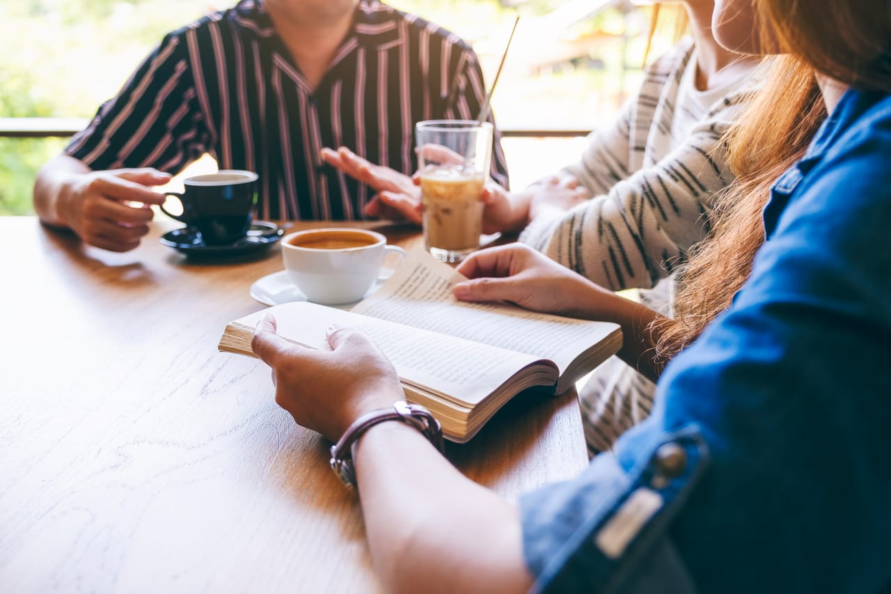 Three people around a cafe table with an open book and coffee between them