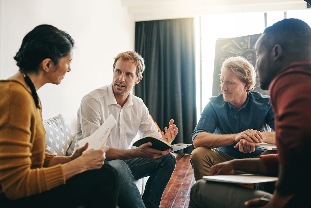 Four people seated in a circle in conversation, one holding an open book