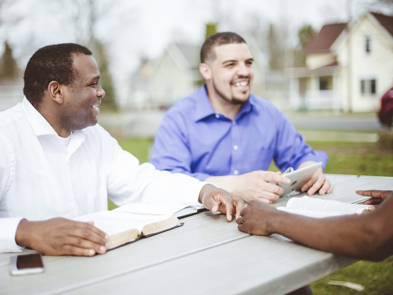 Three men at an outdoor picnic table with open Bibles, talking